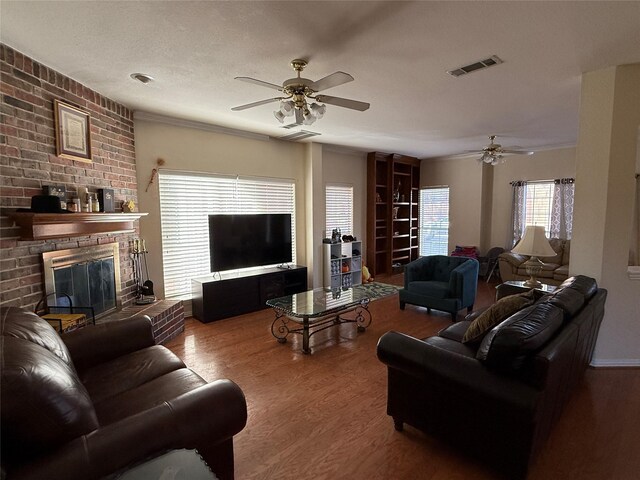 living room with visible vents, a textured ceiling, wood finished floors, a brick fireplace, and ceiling fan