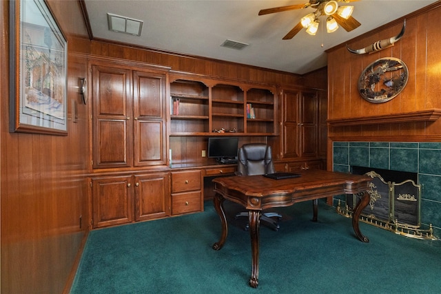 carpeted home office with visible vents, ceiling fan, a tile fireplace, and wooden walls