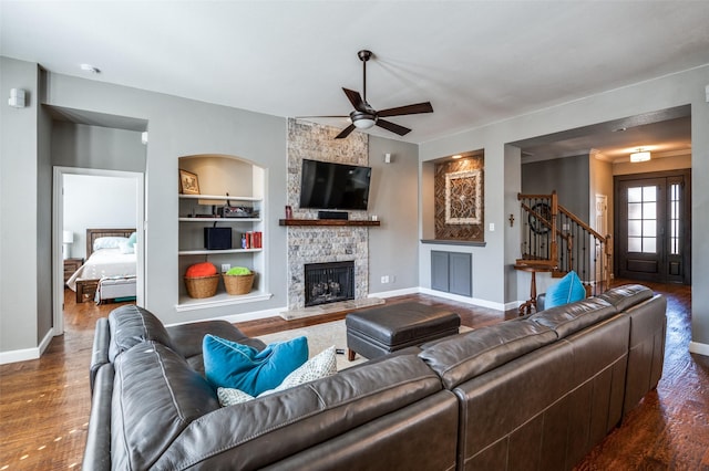 living area with built in shelves, ceiling fan, baseboards, dark wood finished floors, and a stone fireplace