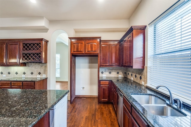 kitchen featuring a sink, stainless steel dishwasher, dark wood-style floors, dark stone counters, and arched walkways