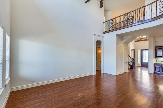 unfurnished living room with stairway, baseboards, arched walkways, dark wood-style flooring, and ceiling fan