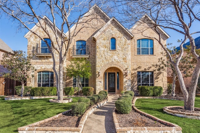 french country home featuring brick siding, stone siding, a front yard, and a balcony