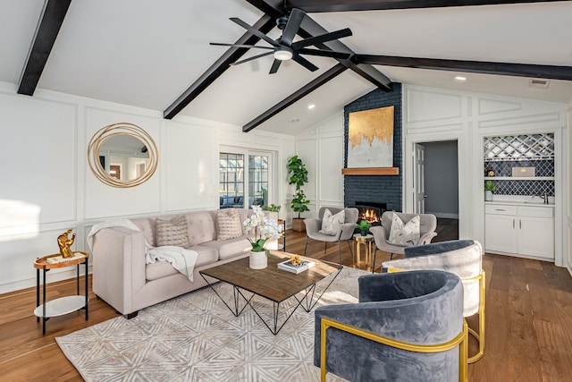 living room with a brick fireplace, ceiling fan, lofted ceiling with beams, wood finished floors, and a decorative wall