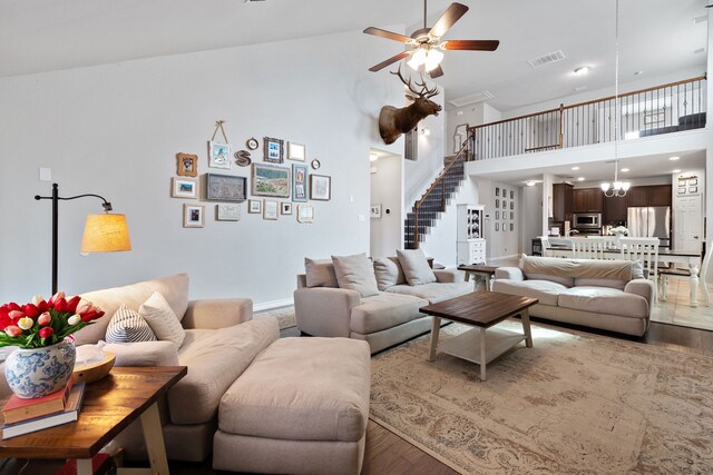 living room with visible vents, high vaulted ceiling, light wood-style flooring, ceiling fan with notable chandelier, and stairway