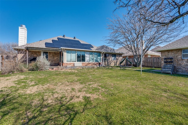 back of house featuring brick siding, fence, roof mounted solar panels, a lawn, and a chimney