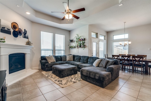 living area featuring a glass covered fireplace, a tray ceiling, light tile patterned floors, and ceiling fan with notable chandelier