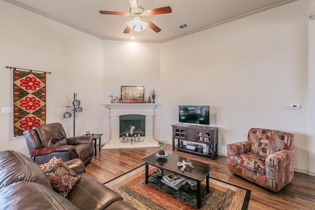 living room with a ceiling fan, wood finished floors, visible vents, a fireplace with raised hearth, and ornamental molding