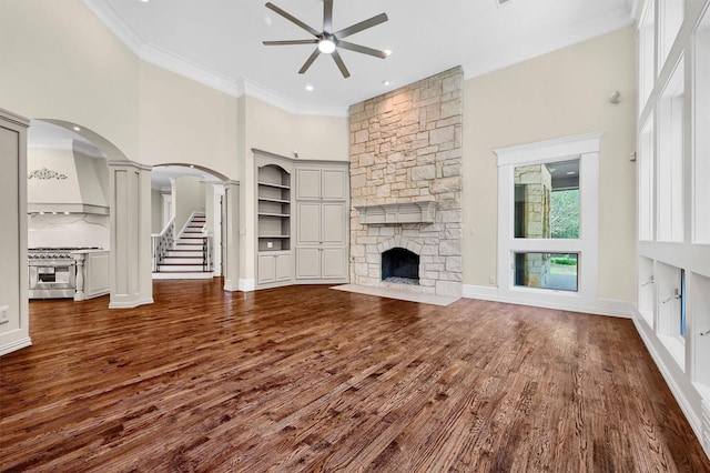 unfurnished living room with ornamental molding, a ceiling fan, arched walkways, a fireplace, and dark wood-style flooring