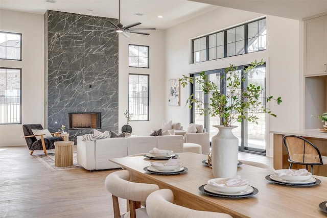 dining room featuring ceiling fan, a premium fireplace, light wood-style flooring, and a towering ceiling