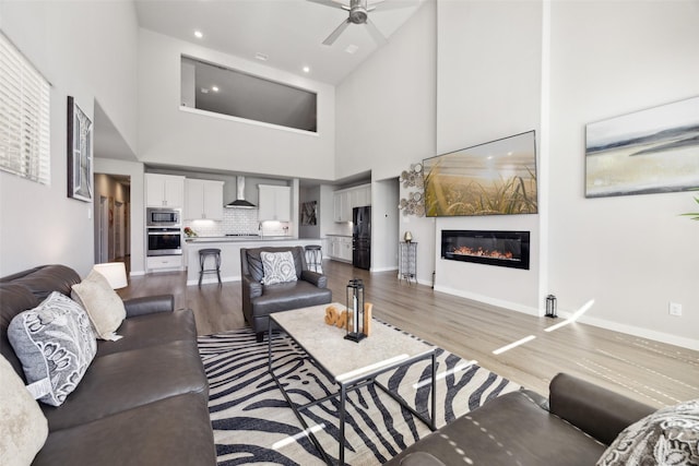 living room with wood finished floors, baseboards, ceiling fan, a towering ceiling, and a glass covered fireplace