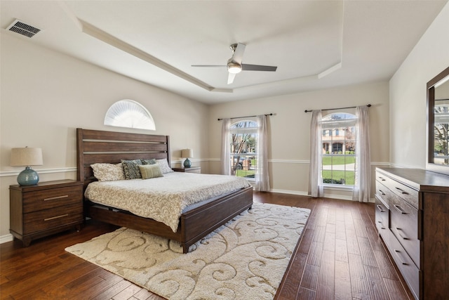 bedroom featuring dark wood-style floors, baseboards, visible vents, a tray ceiling, and ceiling fan