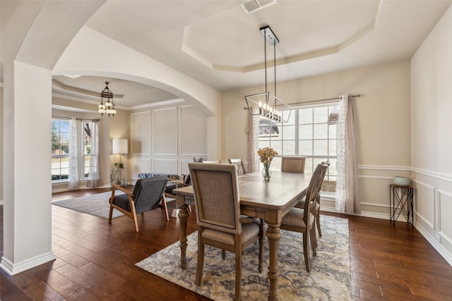 dining area with visible vents, dark wood finished floors, a tray ceiling, arched walkways, and a decorative wall