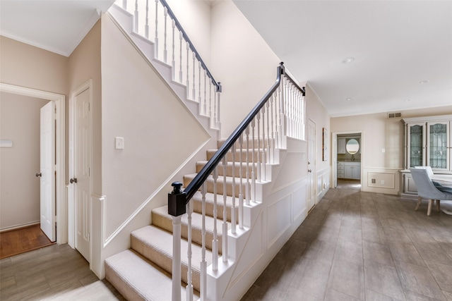 staircase featuring crown molding, a decorative wall, visible vents, and wainscoting