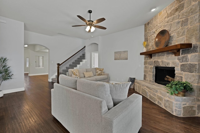 living area featuring stairs, arched walkways, a stone fireplace, and dark wood-style flooring