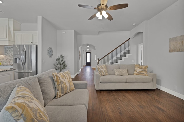 living room featuring dark wood-type flooring, baseboards, stairway, arched walkways, and a ceiling fan