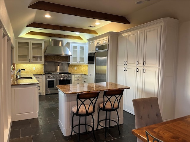 kitchen featuring a kitchen island, a sink, white cabinets, built in appliances, and wall chimney exhaust hood