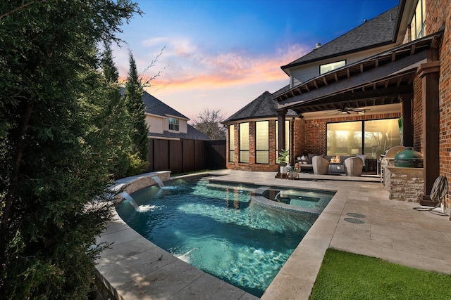 pool at dusk featuring a pool with connected hot tub, fence, exterior kitchen, a patio area, and a ceiling fan