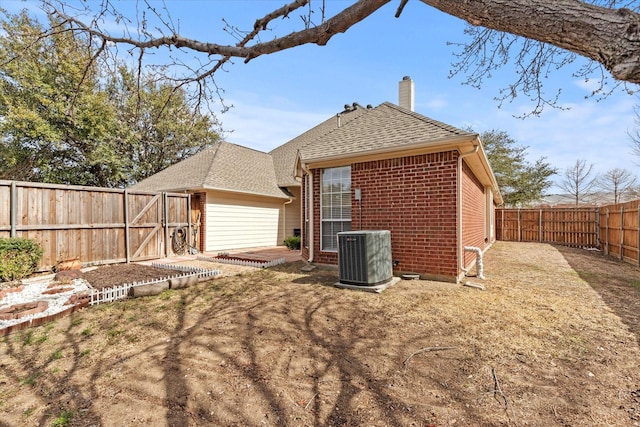 back of property featuring brick siding, central AC, roof with shingles, a chimney, and a fenced backyard