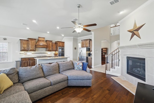 living area featuring visible vents, crown molding, a tiled fireplace, stairway, and dark wood-style flooring