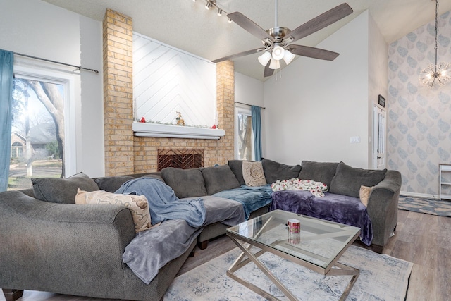 living room featuring wood finished floors, high vaulted ceiling, a textured ceiling, a brick fireplace, and ceiling fan with notable chandelier