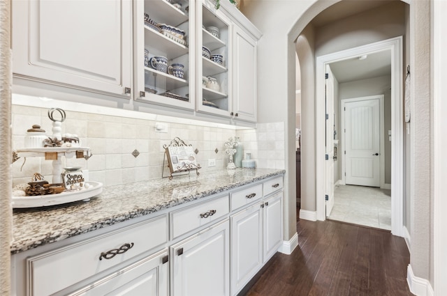 kitchen with dark wood-style floors, light stone countertops, arched walkways, white cabinets, and glass insert cabinets