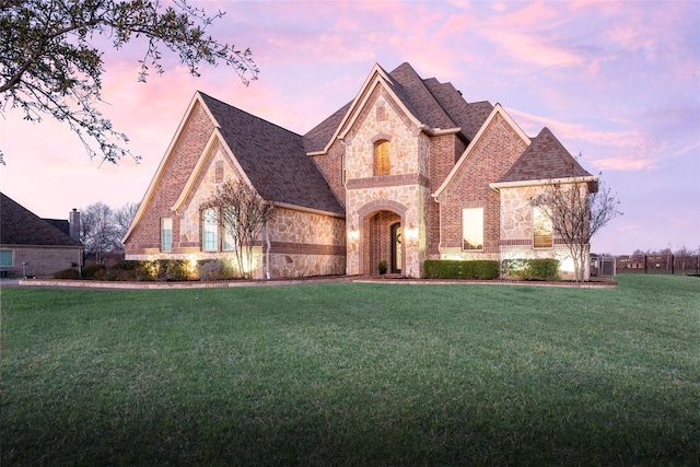 french country inspired facade with brick siding, stone siding, a shingled roof, and a front lawn