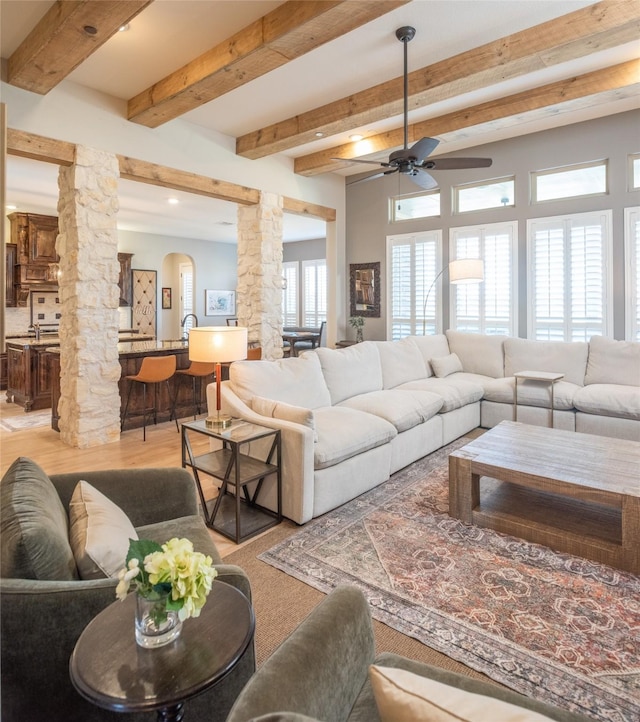 living room featuring beam ceiling, light wood-style flooring, a ceiling fan, arched walkways, and decorative columns