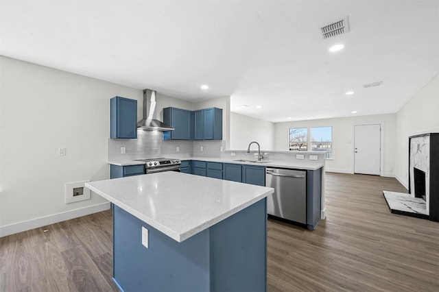 kitchen with wall chimney range hood, a peninsula, stainless steel appliances, blue cabinets, and a sink