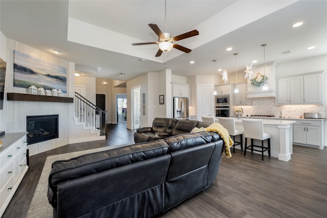 living room with a large fireplace, dark wood finished floors, a tray ceiling, recessed lighting, and a ceiling fan