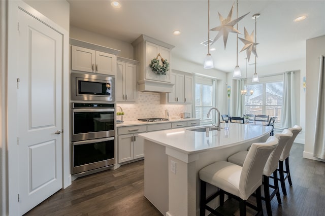 kitchen with a center island with sink, a sink, dark wood-type flooring, appliances with stainless steel finishes, and backsplash