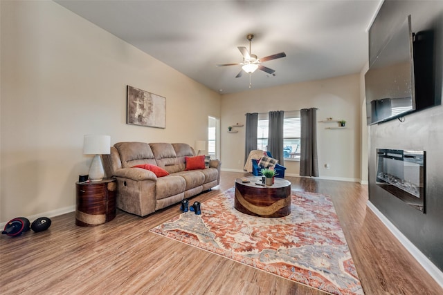 living area with a glass covered fireplace, a ceiling fan, baseboards, and wood finished floors