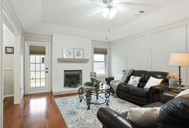 living area featuring visible vents, wood finished floors, a decorative wall, a brick fireplace, and ceiling fan