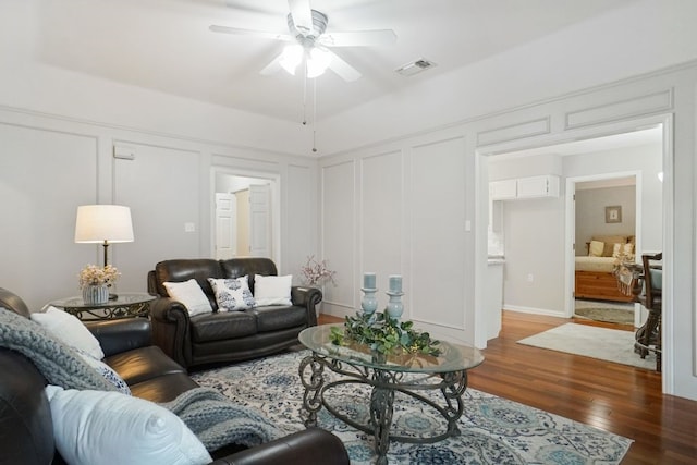 living room with ceiling fan, visible vents, wood finished floors, and a decorative wall