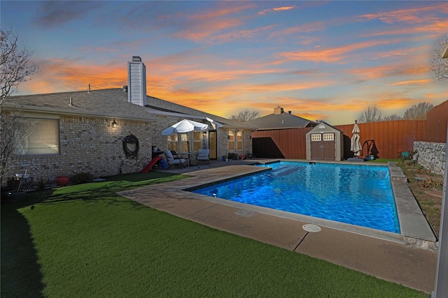 pool at dusk featuring a storage shed, a yard, an outdoor structure, a fenced backyard, and a patio