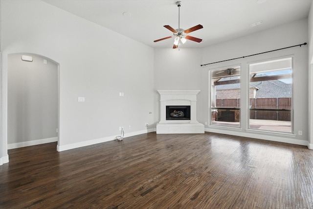 unfurnished living room featuring a fireplace with raised hearth, dark wood-type flooring, baseboards, ceiling fan, and arched walkways