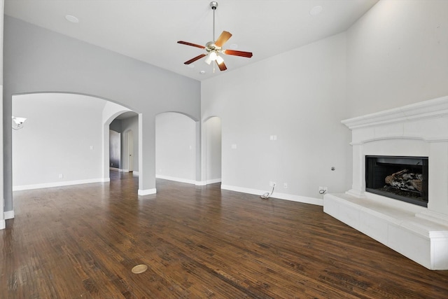 unfurnished living room featuring a fireplace with raised hearth, baseboards, ceiling fan, dark wood finished floors, and arched walkways