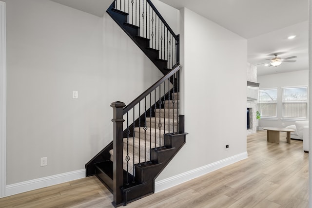 stairway with a ceiling fan, a large fireplace, baseboards, and wood finished floors