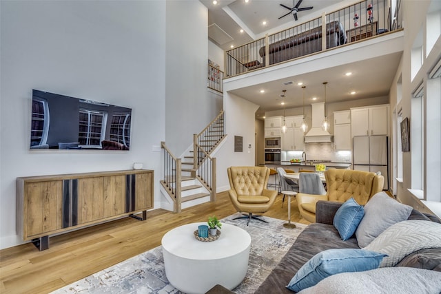 living room with a ceiling fan, baseboards, light wood finished floors, stairs, and a towering ceiling