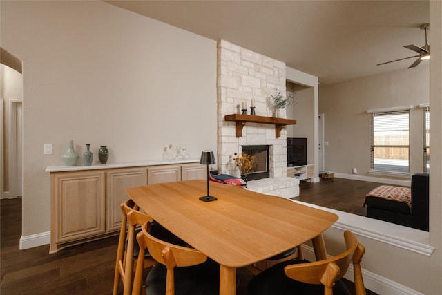 dining space with a stone fireplace, a ceiling fan, dark wood-style flooring, and baseboards