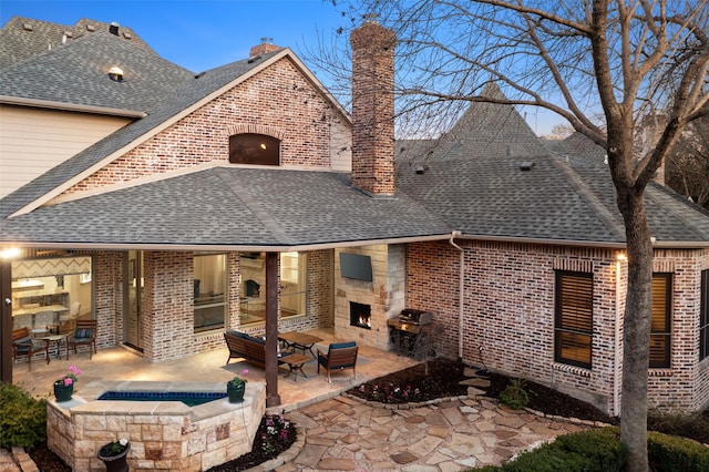 back of house featuring roof with shingles, a chimney, a lit fireplace, a patio area, and brick siding