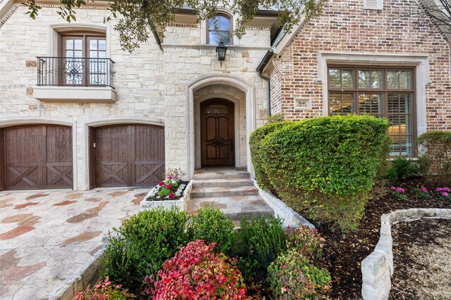 view of exterior entry with a balcony, driveway, an attached garage, stone siding, and brick siding