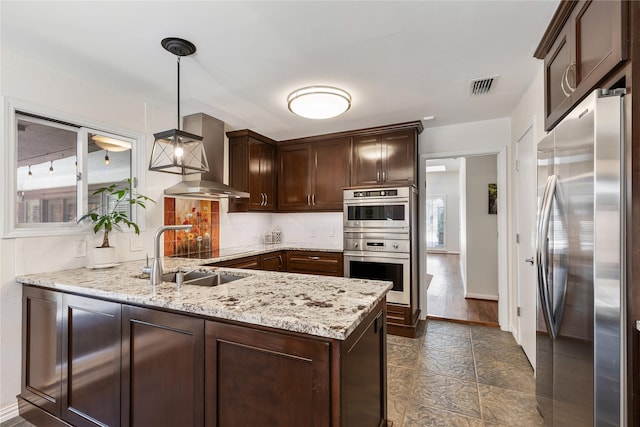 kitchen featuring visible vents, a sink, stainless steel appliances, dark brown cabinetry, and wall chimney range hood