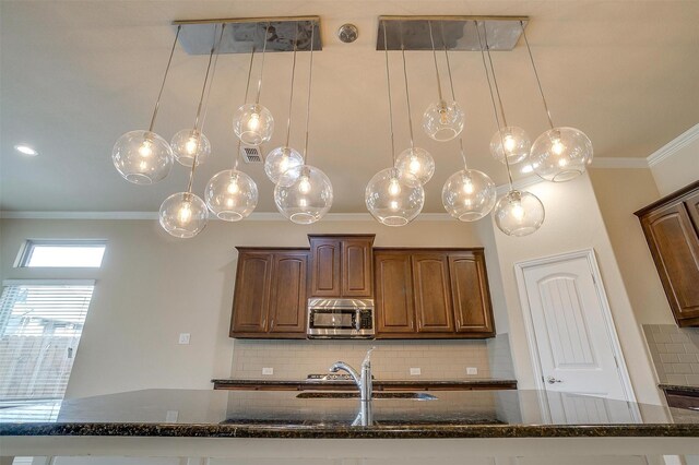 kitchen with a sink, stainless steel microwave, backsplash, dark stone counters, and crown molding