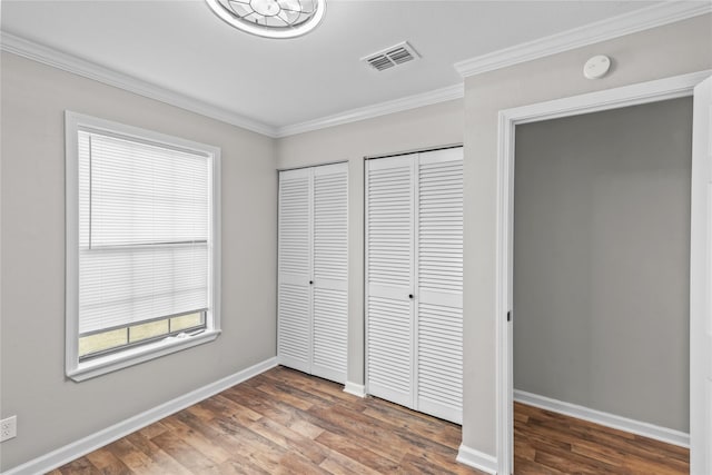 unfurnished bedroom with wood finished floors, baseboards, visible vents, ornamental molding, and multiple closets