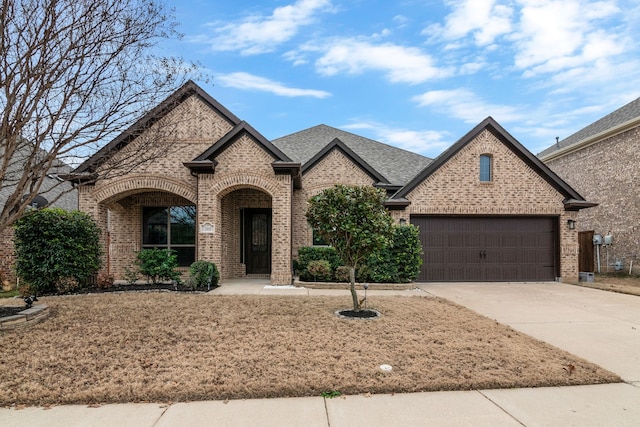 french country inspired facade featuring brick siding, a garage, concrete driveway, and roof with shingles