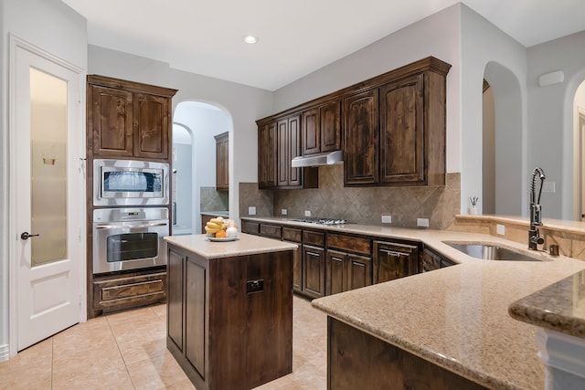 kitchen featuring a sink, stainless steel appliances, dark brown cabinetry, under cabinet range hood, and tasteful backsplash