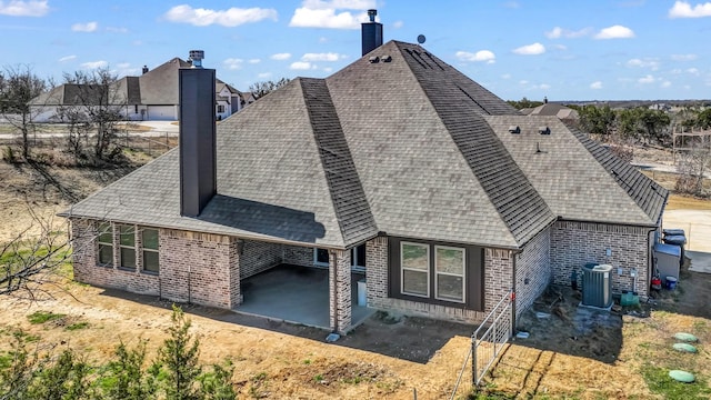 back of property featuring brick siding, central AC unit, a chimney, and a shingled roof