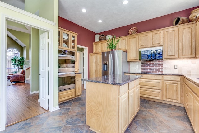 kitchen with dark stone countertops, light brown cabinets, a kitchen island, decorative backsplash, and appliances with stainless steel finishes