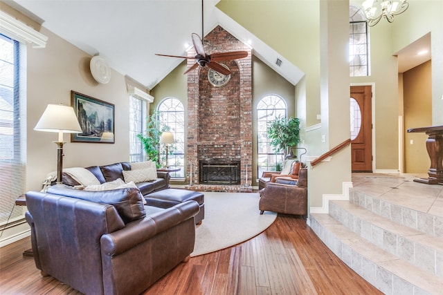 living area featuring stairway, wood finished floors, high vaulted ceiling, a fireplace, and ceiling fan with notable chandelier