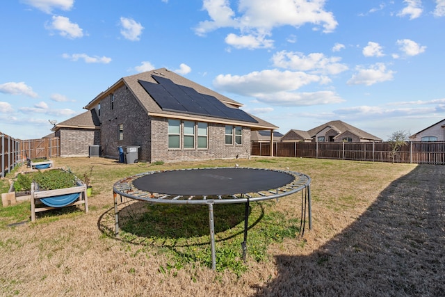 back of house with a fenced backyard, a trampoline, a lawn, brick siding, and roof mounted solar panels